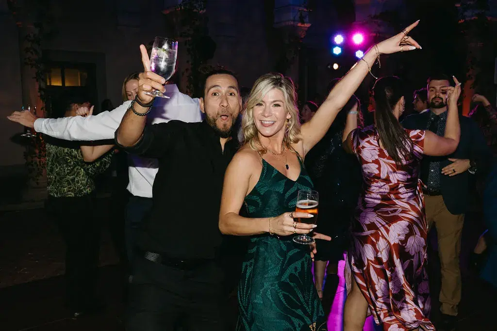 Two guests posing playfully with drinks raised on a wedding dance floor lit in purple