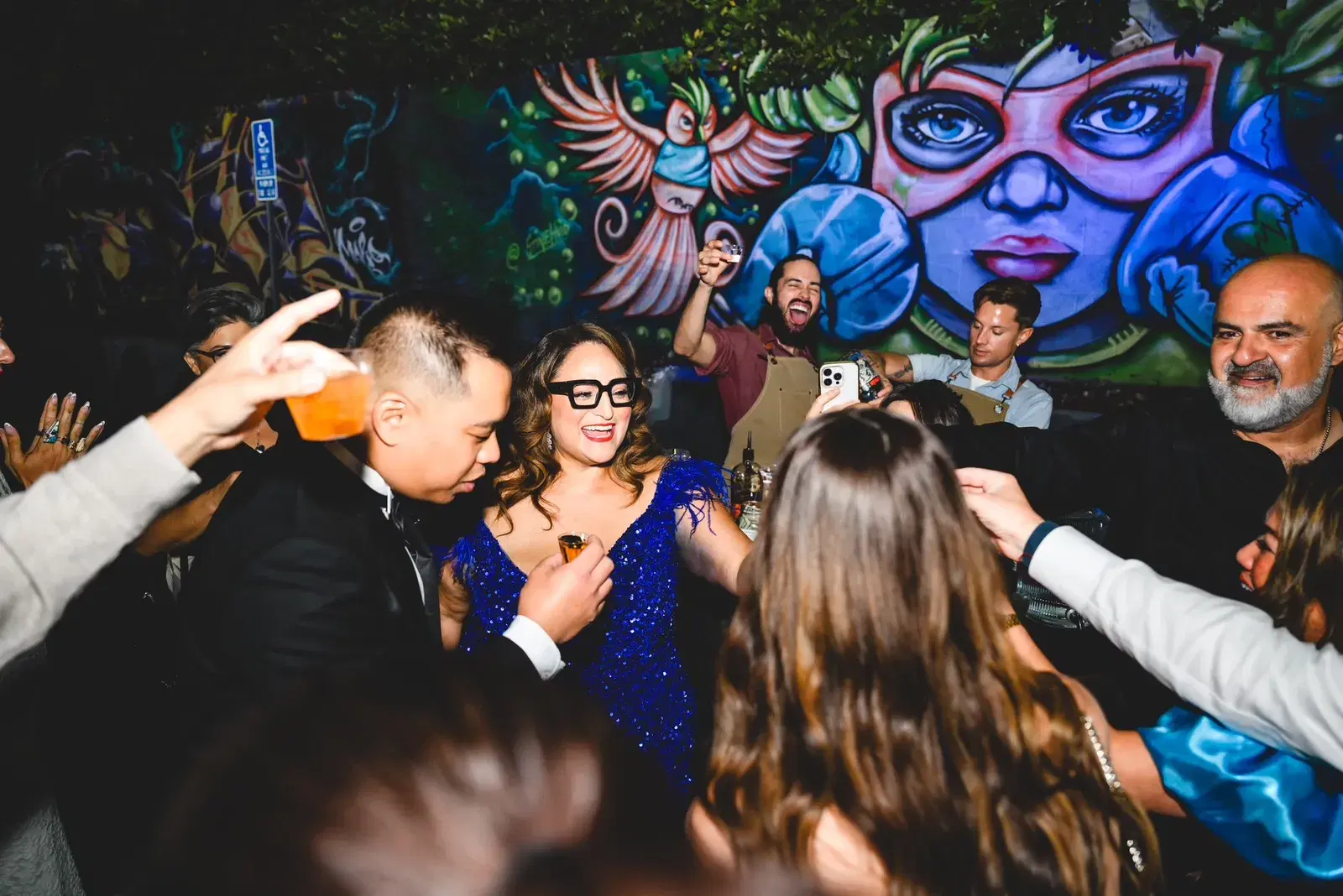 Guest of honor in a blue sequin dress laughing with friends at an outdoor party in front of a colorful mural