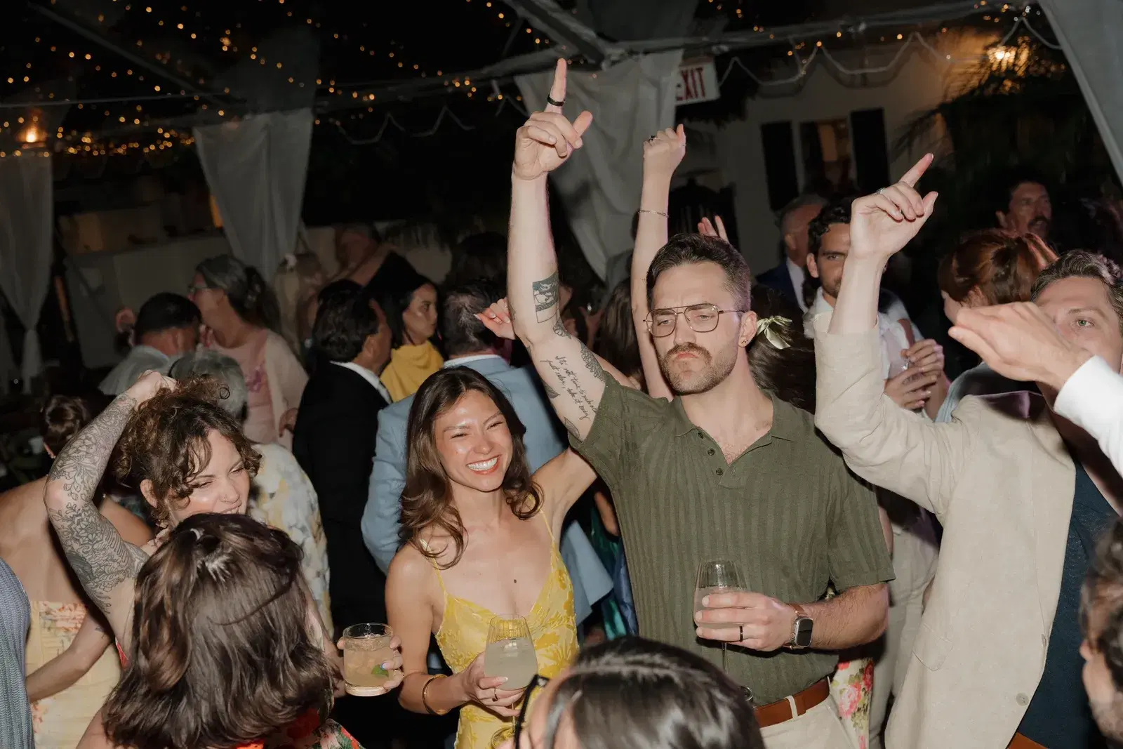 Guests dancing with hands in the air under a tent strung with lights at a wedding reception