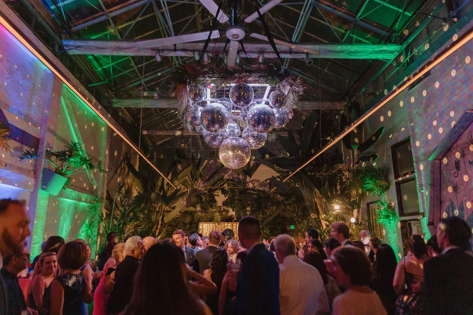 Cluster of mirrored disco balls suspended above a crowded dance floor at a tropical greenhouse-style event venue