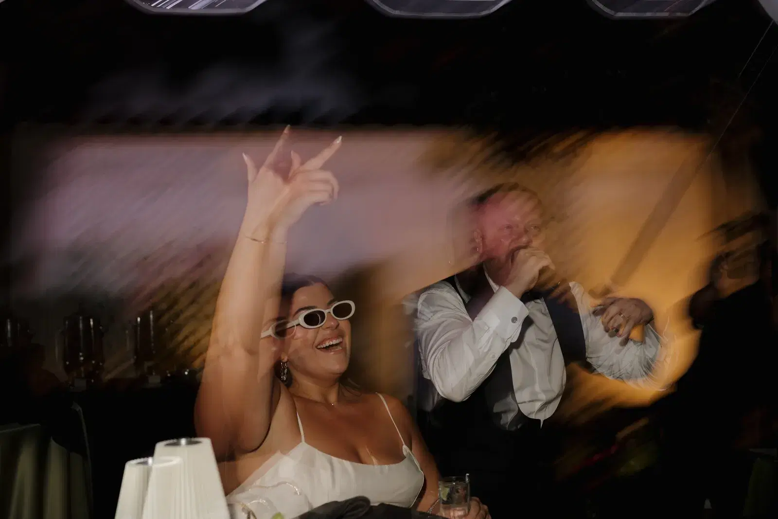 Bride in white sunglasses laughing and dancing during a high-energy wedding reception dance party