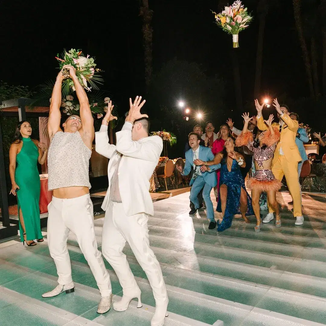 Two grooms tossing bouquets to cheering guests on a poolside dance floor at a Los Angeles wedding reception