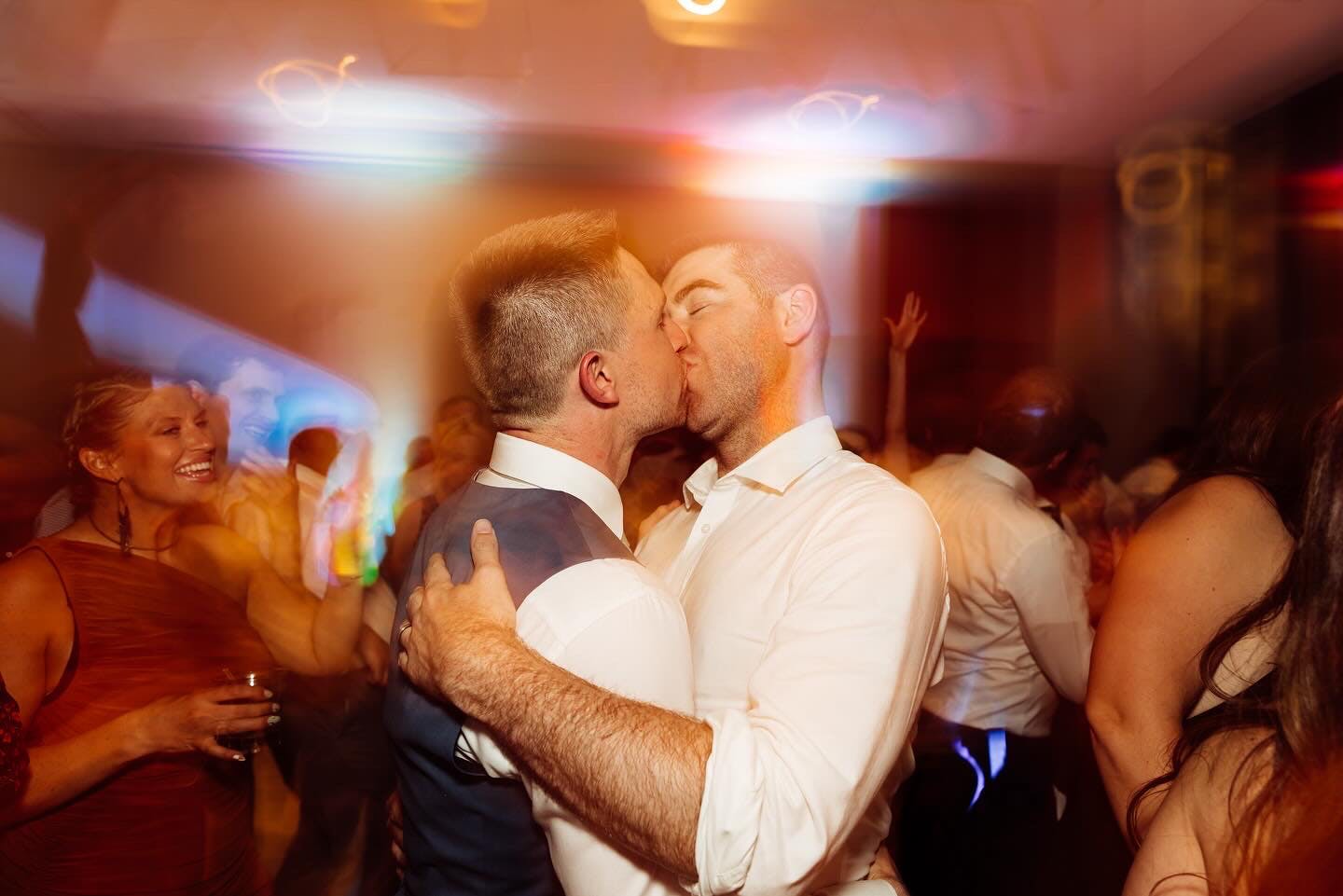 Two grooms embrace and kiss on the dance floor while guests smile and cheer around them